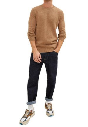 Homme portant un jean en denim bleu foncé retroussé, un pull beige à taches, des chaussettes blanches et des baskets multicolores, se tenant avec une main dans la poche.
