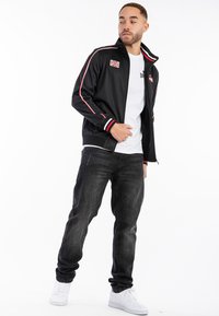 Svart zip-up jacka med röda och vita ränder, brittisk flagga patch, bärs över en vit t-shirt, ihop med mörka jeans och vita sneakers.