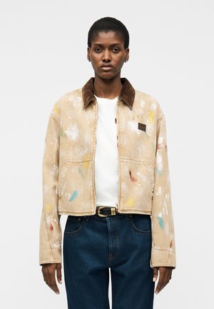 Personne portant une veste courte beige avec des éclaboussures de peinture, un col marron, une chemise blanche, un jean bleu foncé et une ceinture noire sur un fond uni.