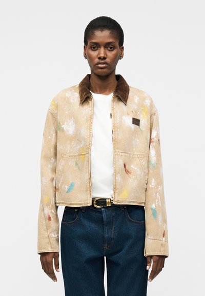 Personne portant une veste courte beige avec des éclaboussures de peinture, un col marron, une chemise blanche, un jean bleu foncé et une ceinture noire sur un fond uni.