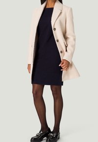 Cappotto di colore crema, dalla trama, con colletto a punte, chiusura a bottoni e tasche laterali, indossato sopra un vestito in maglia scuro e scarpe nere.
