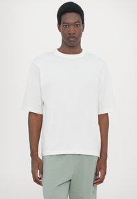 Bijela pamučna majica kratkih rukava, okruglog izreza i opuštenog kroja. Tekstuirani materijal, u kombinaciji s svijetlozelenim hlačama.