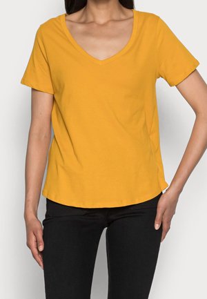 Żółta bawełniana koszulka z dekoltem w serek, krótkimi rękawami i prostym dołem. Połączona z czarnymi dżinsami, prezentowana na jednolitym tle.