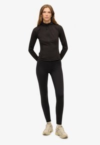 Czarna sportowa kurtka z wysokim kołnierzem i zapięciem na zamek, w połączeniu z czarnymi legginsami i beżowymi trampkami. Gładki, dopasowany krój.