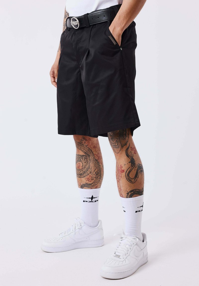 Homme portant un short noir, une ceinture noire, des chaussettes blanches avec un motif noir, des baskets blanches et des jambes tatouées sur un fond uni.