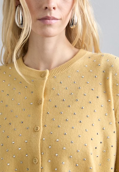 Κίτρινη πλεκτή ζακέτα με στρογγυλή λαιμόκοψη, διακοσμημένη με ασημένια κρυστάλλινα στρας και πέντε κουμπιά μπροστά. Λεια υφή.