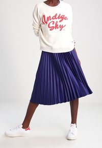 Sweat-shirt crème avec imprimé "Indigo Sky" en rouge, associé à une jupe midi plissée bleu foncé. Des baskets blanches avec des touches rouges complètent le look.