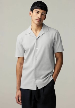 Camicia a maniche corte, di colore grigio chiaro, con colletto e sottili righe verticali, dotata di sei bottoni. Indossata con pantaloni scuri, dalla vestibilità rilassata.