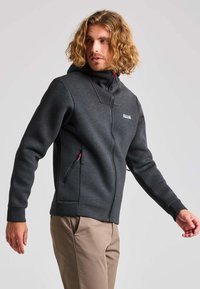 Donkergrijze ritsvest met hoge kraag, zijzakken en rode ritsdetails. Gemaakt van zachte stof met een aansluitend ontwerp.