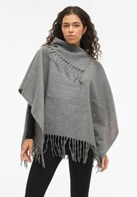 Sieviete ar cirtainiem matiem, uzvilkusi pelēku vilnas pončo ar malu detaļām, virs melna pielīdzena krekla ar garām piedurknēm un melnām biksēm, stāv uz baltas fona.