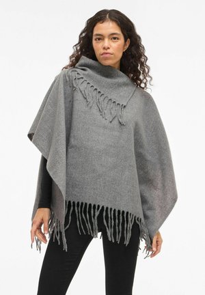 Kvinde med krøllet hår iført en grå frynset uldponcho over en sort langærmet bluse og sorte bukser, som står foran en hvid baggrund.