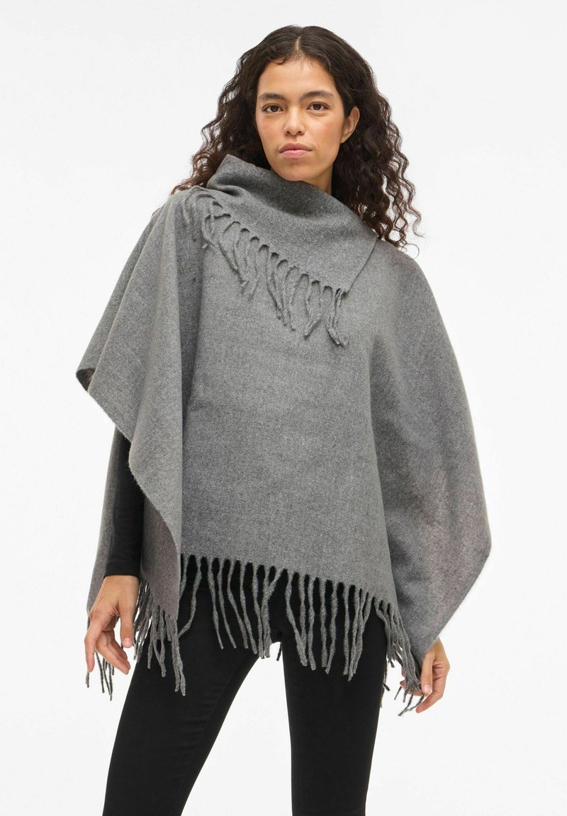 Sieviete ar cirtainiem matiem, uzvilkusi pelēku vilnas pončo ar malu detaļām, virs melna pielīdzena krekla ar garām piedurknēm un melnām biksēm, stāv uz baltas fona.