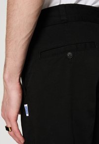 Le bras gauche avec une bague repose à côté d'un pantalon noir comprenant une poche arrière avec un bouton et une petite étiquette bleue "HUGO".