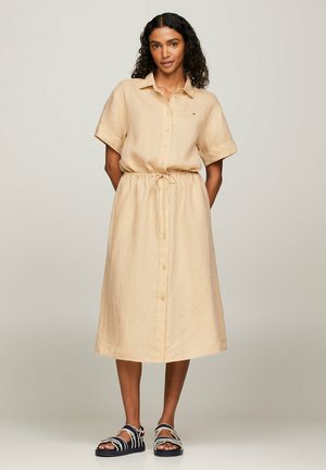 Abito camicia in lino beige con maniche corte, chiusura a bottoni davanti, vita regolabile con cordino e orlo fino al ginocchio, abbinato a sandali neri.