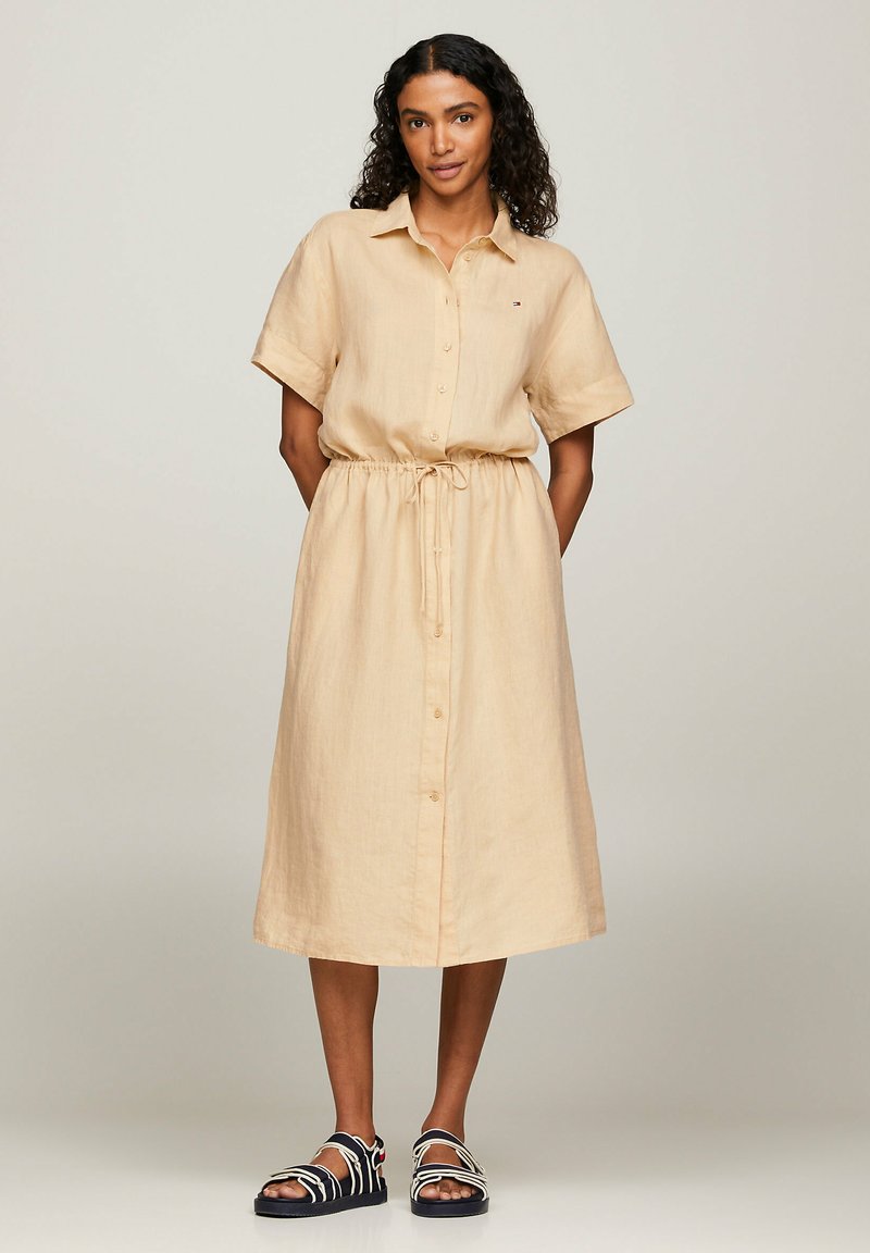 Abito camicia in lino beige con maniche corte, chiusura a bottoni davanti, vita regolabile con cordino e orlo fino al ginocchio, abbinato a sandali neri.