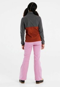 Pull en polaire gris et rouille, avec un col montant et des manches longues. Associé à un pantalon évasé rose et des bottes noires. Design simple avec des caractéristiques.