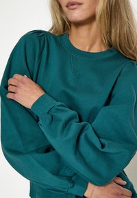 Felpa color teal con collo a giro, maniche a sbuffo e polsini a coste. Realizzata in un tessuto morbido con una texture liscia. Design semplice.