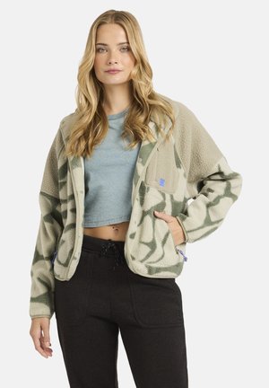 Camouflage-Fleecejacke in Grün und Beige, mit lockerer Passform, Druckknöpfen und Seitentaschen. Kombiniert mit einem kurz geschnittenen blauen Oberteil und dunklen Hosen.