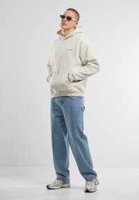 Lys grå hoodie med en forlommer og et blåt logo, parret med blå løse jeans og sølvfarvede løbesko. Slanke solbriller fuldender looket.