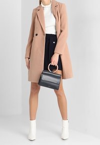 Manteau rose clair avec un design ajusté, top blanc à côtes, shorts taille haute navy, sac à main noir avec une poignée en bois, et bottines blanches.