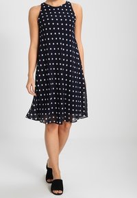 Mörkblå ärmlös klänning med vita prickar, utställd silhuett och genomskinlig nederkant. Bärs med svarta öppna skor.
