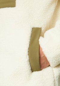 Hand in olijfgroen afgezakte zak van witte textuurrijke fleecejas met zachte, pluizige stof en zichtbare naaddetails.