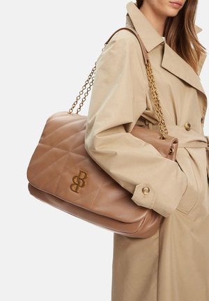 Frau in beigem Trenchcoat, die eine große, gesteppten beige Lederhandtasche mit goldenen Kettenriemen und Doppel-B-Logo trägt.