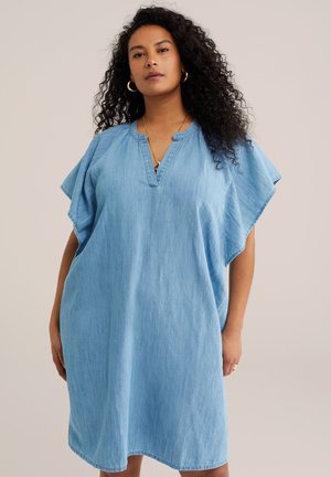 Garbanotomis plaukomis moteris dėvi laisvą šviesiai mėlynos spalvos džinsinę suknelę su trumpomis plazdančiomis rankovėmis ir V formos iškirpte, stovinti priešais paprastą foną.