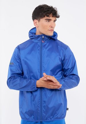 Mann, der eine leuchtend blaue Kapuzen-Windjacke trägt, steht mit verschränkten Händen und schaut nach rechts auf weißem Hintergrund.