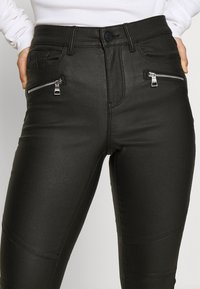 Pantalon slim en simili cuir noir avec poches avant zippées, coupe ajustée et détails de surpiqûres le long des jambes. Texture lisse et finition brillante.