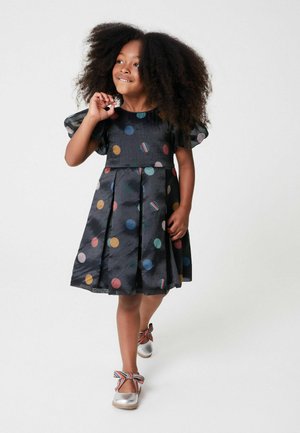 Schwarzes Kleid mit mehrfarbigen Punkten, kurzen Puffärmeln und gefälteltem Rock. Silberschuhe mit gestreiften Bandakzenten.