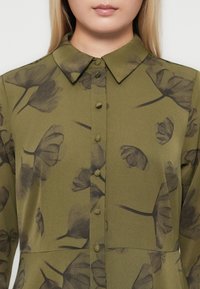 Primer piano di una persona che indossa una camicia a bottoni verde oliva con un motivo floreale e un colletto a punta.