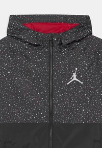 Chaqueta negra con capucha, con un patrón de salpicaduras en blanco y rojo. Cierre frontal con cremallera y un logo blanco en el pecho. Textura suave.