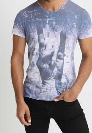 Homme portant un t-shirt bleu et blanc avec une grande empreinte de main faisant le signe du rock and roll à l'avant, associé à un pantalon noir.