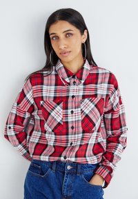 Chemise à carreaux rouge et noir avec deux poches avant, fermeture à boutons et col arrondi, associée à un jean en denim bleu.