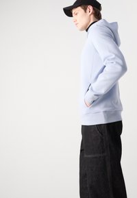 Svijetloplava hoodie napravljena od mekog materijala, s kenguruvim džepom i rebrastim rubom. Uparen s tamnoplavim trapericama i crnom kapom.