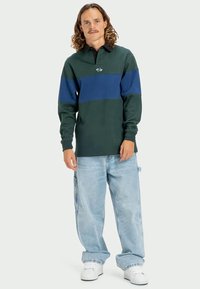 Zelené polo tričko s modrým horizontálnym pruhom, obsahujúce logo. V kombinácii s svetlomodrými voľnými džínsami a bielymi teniskami.