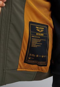Softshell jakna s vanjštinom u zelenoj boji, narančastom mrežastom podstavom i crnim unutrašnjim dijelom. Sadrži oznaku s specifikacijama proizvoda i uputama za njegu.