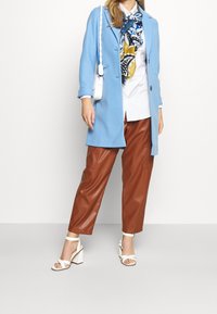 Cappotto azzurro chiaro, camicia bianca, pantaloni in pelle sintetica marrone e sandali con tacco blocco bianchi. Presenta una sciarpa a fantasia e una piccola borsa bianca.