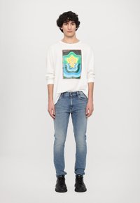 Witte long-sleeve shirt met een veelkleurige geometrische print, gecombineerd met lichtblauwe spijkerbroek en zwarte schoenen, staand tegen een effen achtergrond.