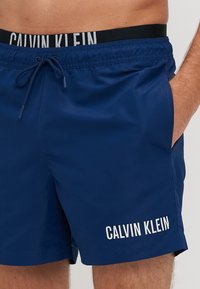 Calções de banho azul-marinho com uma cintura preta apresentando o texto branco "CALVIN KLEIN". Feitos de um tecido suave e leve, com bolsos laterais.