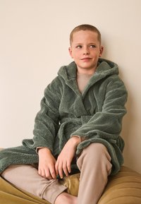 Green, fluffy bathrobe with a hood, belt, and deep pockets, paired with a beige long-sleeve top and pants, seated on a tan cushion.