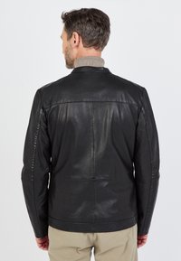 Man in een zwart leren jasje en beige broek, met zijn rug naar de camera gekeerd tegen een effen lichte achtergrond.