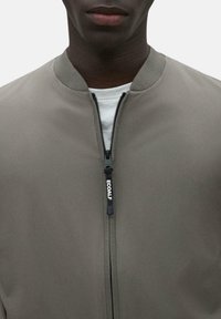 Chaqueta bomber gris con un cuello redondeado, cremallera frontal completa y un logo en la cremallera. Textura de tela lisa y mate. Camiseta blanca lisa debajo.