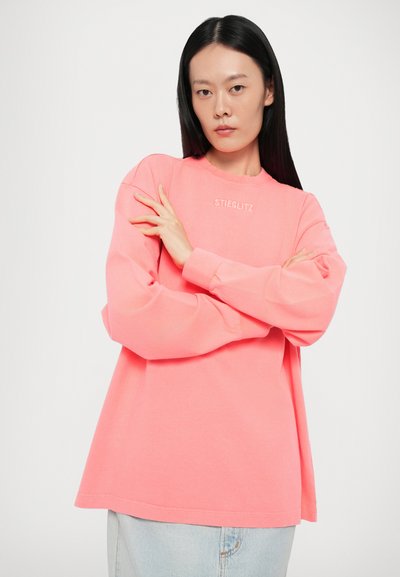 Pink sweatshirt with a crew neckline and long sleeves, features the "STIEGLITZ" logo in white across the chest, worn with light denim jeans.