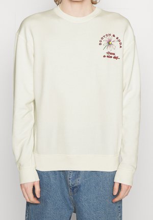 Kremowa bluza z długim rękawem z małym stokrotkowym kwiatem i czerwonym napisem "Scotch & Soda Have a nice day" na górnej lewej stronie klatki piersiowej, noszona z niebieskimi dżinsami.