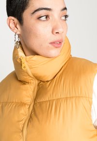 Colete amarelo almofadado com gola alta, fecho frontal e bolsos laterais. Apresenta um tecido liso e brilhante, além de costuras decorativas.