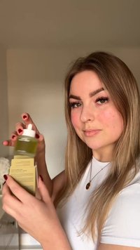 Femme aux longs cheveux tenant un flacon compte-gouttes de liquide vert et une boîte beige, portant une chemise blanche et un collier à l'intérieur.