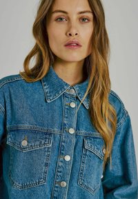 Zila džinsu jaka ar apkakli, priekšējām kabatām un sudraba pogām. Audums izskat�ās teksturēts ar nelielu izbalējumu un redzamām dūrēm.