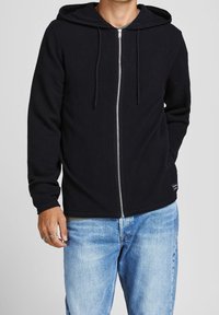 Black zip-up hoodie made of textured fabric, featuring a drawstring hood, front pockets, and a smooth zipper. Paired with light blue jeans.